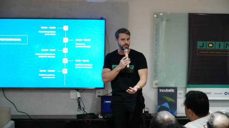 Gabriel Melo apresenta o cronograma do programa JoInT+ para startups, com tela digital ao fundo.